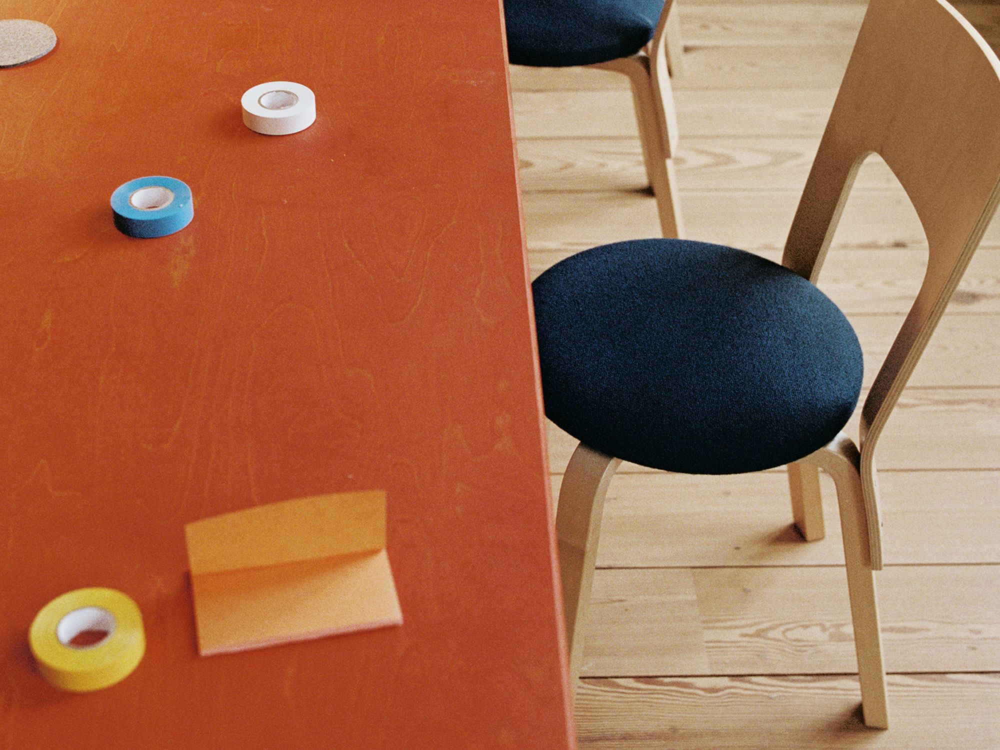 Photograph of a tabel top with tape roles in various colors. Next to the table is a wooden chair with a blue pillow on a wooden floor.